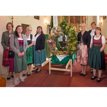 Eine Gruppe von Frauen in traditioneller Kleidung steht um einen Blumenkranz in einer Kirche, mit einem Teppichboden und religiösen Symbolen im Hintergrund.