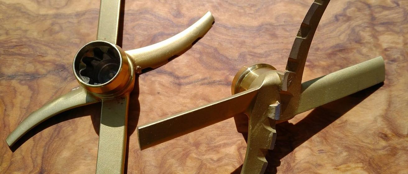 Zwei goldene mechanische Arme liegen auf einer Holzoberfläche. Ein Arm hat eine kreisförmige Öffnung und ein Getriebe im Inneren, während der andere Arm ein blattförmiges Ende hat.