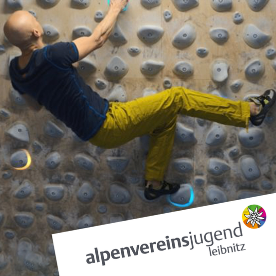 Ein Mann klettert mit einem Lächeln an einer Boulderwand, trägt ein blaues T-Shirt und gelbe Hosen.