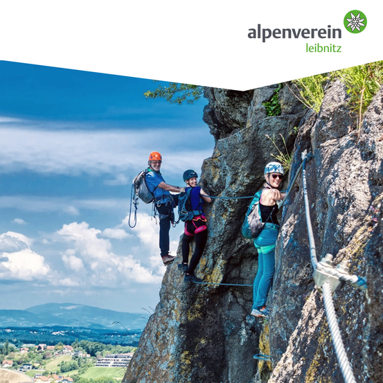 Drei Personen klettern am Fels und tragen Sicherheitsausrüstung. Eine steht vorne, zwei dahinter, verbunden durch Seile. Im Hintergrund sieht man ein Tal mit Bergen und Häusern.