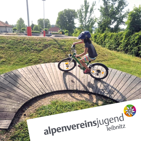 Ein Kind fährt auf einem geschwungenen Holzweg mit dem Fahrrad. Grüner Rasen und Bäume umgeben den Bereich. Unten ist ein Logo zu sehen.
