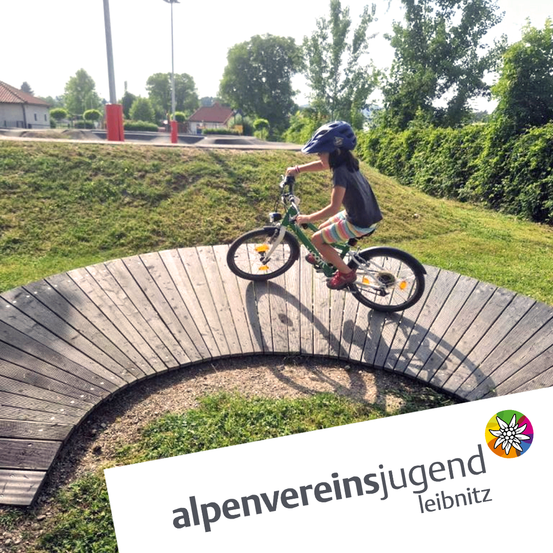 Ein Kind fährt auf einem geschwungenen Holzweg mit dem Fahrrad. Grüner Rasen und Bäume umgeben den Bereich. Unten ist ein Logo zu sehen.