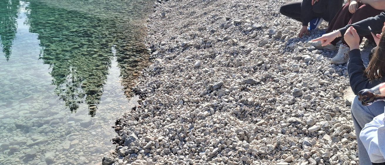 Drei Personen sitzen auf Felsen am Wasser. Eine schreibt, eine sieht sie an und eine beobachtet das Wasser.