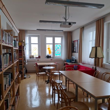 Eine leere Bibliothek mit mehreren Tischen und Stühlen, einem roten Sofa und Regalen voller Bücher. Zwei Fenster haben farbenfrohe Designs.