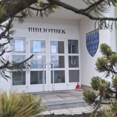 Ein Bibliothekseingang mit Glastüren und einem Schild, das 'BIBLIOTHEK' steht. Rechts befindet sich ein roter Besen und ein Wappen an der Wand.