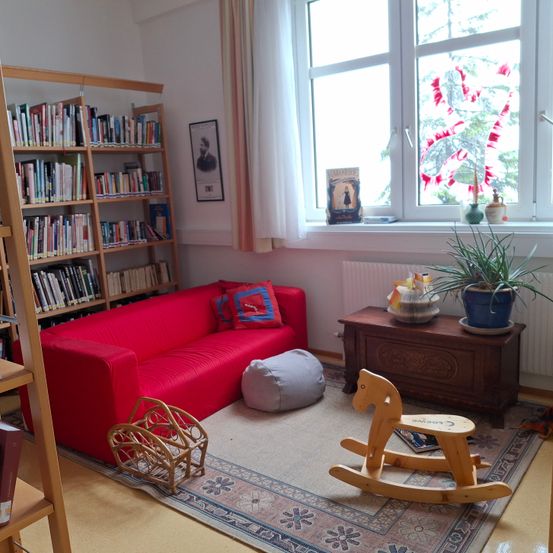 Ein Wohnzimmer mit einem roten Sofa, einem beigen Teppich und einem Holzpferdchen. Ein Bücherregal mit Büchern und einem Bilderrahmen ist links. Ein Fenster mit weißen Vorhängen hat eine Topfpflanze und ein Bild. Ein hölzerner Schrank mit einer Schüssel und einer Vase ist rechts.