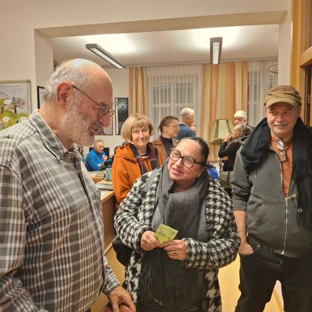 Mehrere Erwachsene in einem Raum, eine Frau hält Geld. Ein Mann in einem karierten Hemd lächelt und ein anderer Mann trägt einen Hut. Andere Leute stehen hinter ihnen und eine Lampe steht auf einem Tisch.