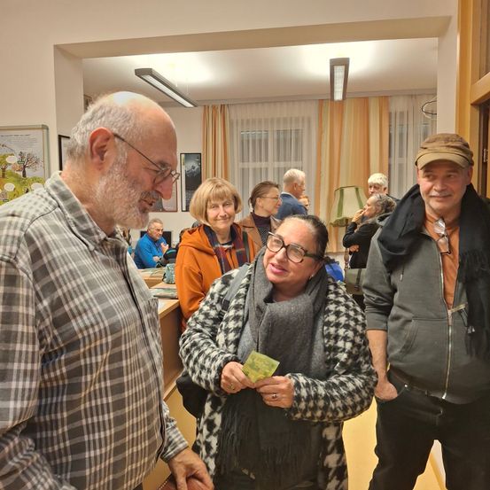 Mehrere Erwachsene in einem Raum, eine Frau hält Geld. Ein Mann in einem karierten Hemd lächelt und ein anderer Mann trägt einen Hut. Andere Leute stehen hinter ihnen und eine Lampe steht auf einem Tisch.