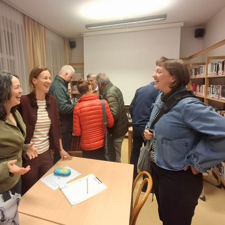Eine Gruppe Erwachsener trifft sich in einer Bibliothek, in Gespräche vertieft. Ein Tisch im Vordergrund zeigt verschiedene Gegenstände, und Regale mit Büchern säumen die Wände.