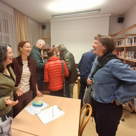 Eine Gruppe Erwachsener trifft sich in einer Bibliothek, in Gespräche vertieft. Ein Tisch im Vordergrund zeigt verschiedene Gegenstände, und Regale mit Büchern säumen die Wände.