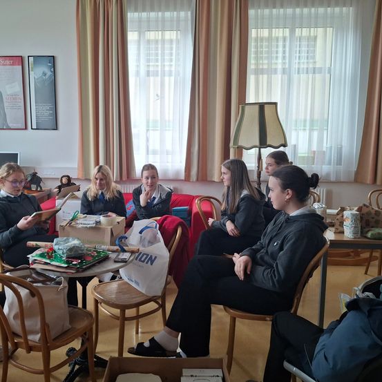 Eine Gruppe von Frauen sitzt in einem Raum um einen Tisch herum, liest, redet und schaut sich Gegenstände an. Einige Kisten und Taschen sind auf dem Tisch und Boden. Fenster mit Vorhängen und einer Lampe befinden sich hinter ihnen.