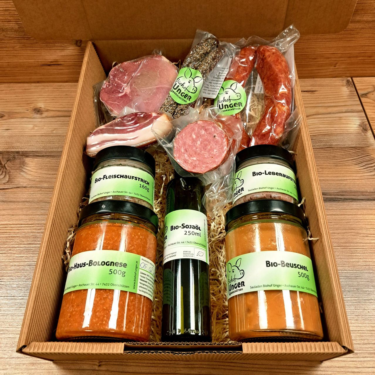 Eine Schachtel mit verschiedenen Fleischprodukten, darunter Würste, Salami und Rinderscheiben. Es gibt auch Gläser mit Sauce und eine Flasche Öl. Alle Artikel sind biologisch.
