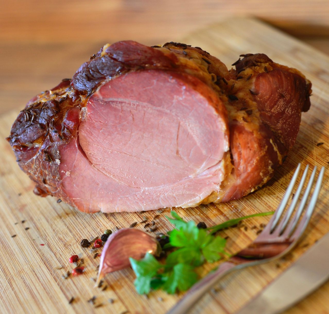 Ein geschnittener Schinkenbraten mit knuspriger Kruste liegt auf einem Holzschneidbrett. Daneben befinden sich eine Gabel, ein Messer und einige Kräuter und Gewürze.