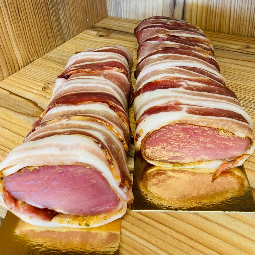 Zwei in Speck gewickelte Würste liegen auf einem Holztisch, einer auf einem goldenen Teller, mit einem goldenen Reflex darunter.