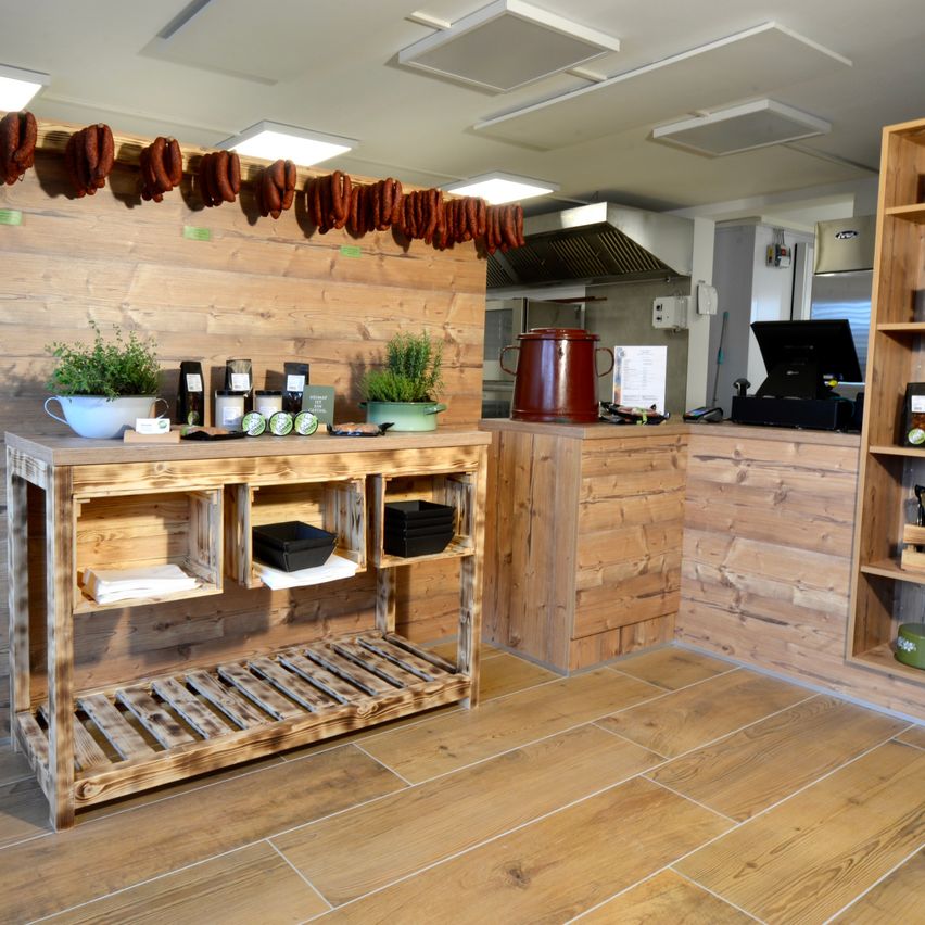 Ein Holztresen und Regale mit Pflanzen und Behältern stehen in einer Küche. Würste hängen an der Wand.