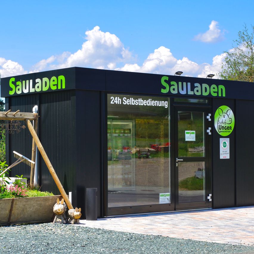 Ein schwarzes Gebäude mit dem Schild 'SauLaden' und einer Tür, die '24h Selbstbedienung' besagt. Es hat ein grünes Logo mit dem Text 'Unser' und einem Schwein. Ein Holzstab und ein Topf stehen neben dem Gebäude. Der Boden ist mit Kies bedeckt.