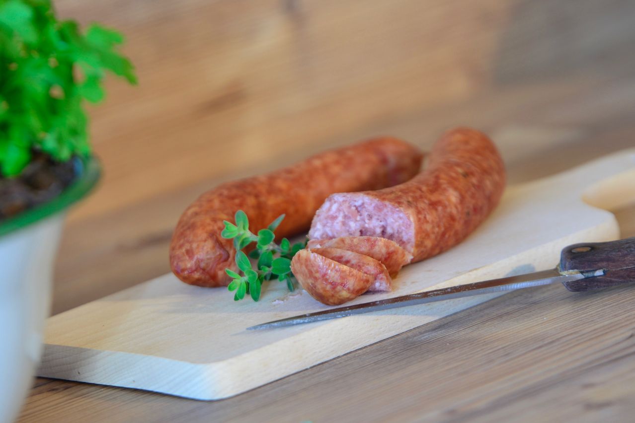 Zwei Würste auf einem Holzbrett, teilweise geschnitten mit einem Messer und begleitet von frischen Kräutern auf einer Holzoberfläche.