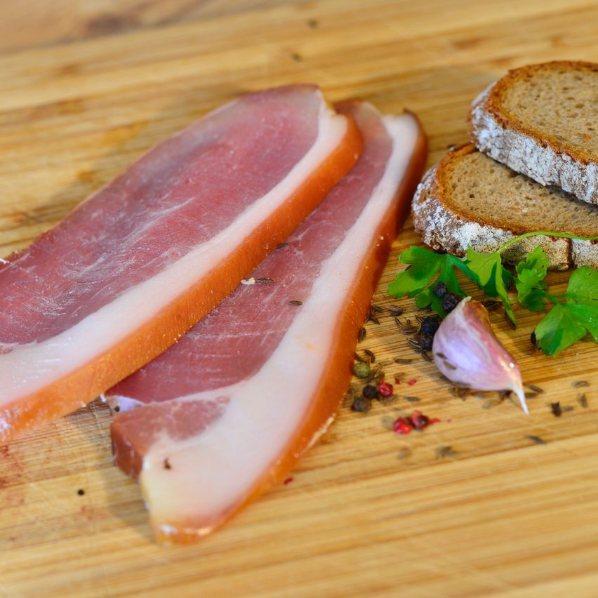 Zwei Scheiben Speck liegen auf einem Holzschneidebrett mit einer Scheibe Brot, Petersilie und Knoblauchzehen.