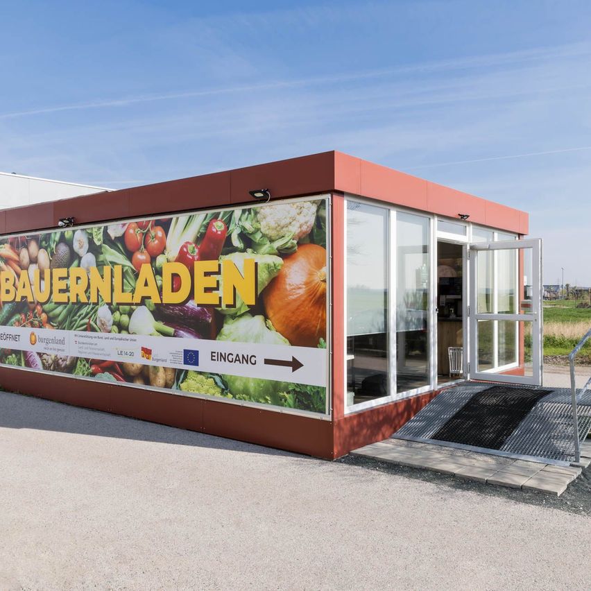 Ein rotes Gebäude mit einer Glasfassade zeigt ein großes Poster von Gemüse und Obst. Der Text 'Bauernladen' ist prominent angebracht, mit einem Pfeil, der auf den Eingang zeigt. Eine Rampe führt zum Eingang.