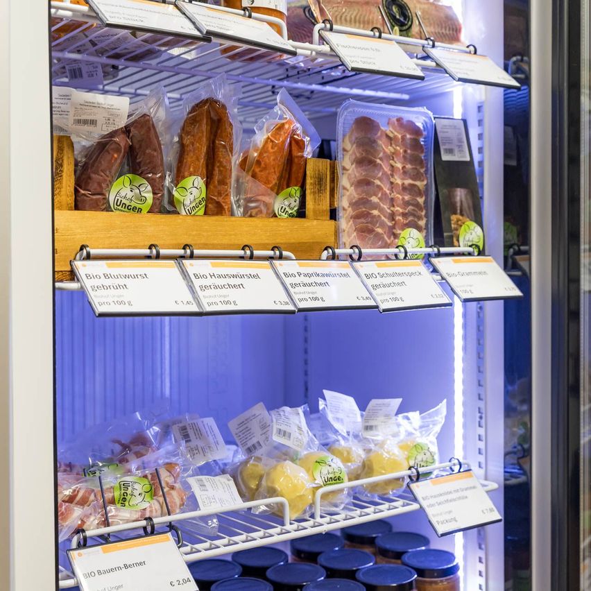 Eine Kühlvitrine mit verschiedenen Packungen Bio-Fleisch, einschließlich Bio-Blutwurst, Bio-Hauswurst, Bio-Paprikawurst und Bio-Schulterspeck, alles unter blauem Licht.