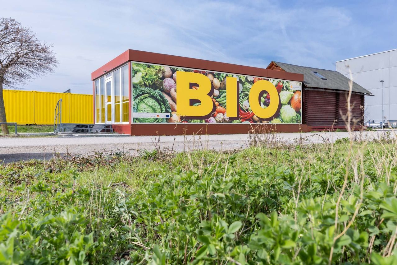 Ein Gebäude mit einem großen gelben Bio-Schild an einem sonnigen Tag, mit grünem Gras und einem gelben Container im Hintergrund.