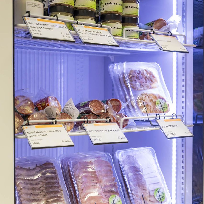 Ein offener Kühlschrank mit verschiedenen Lebensmitteln. Die oberste Ablage enthält Gläser mit Bio Schweineschmalz, Bio Austrichfleisch und Bio Grammelwurst. Die untere Ablage hat Packungen von Bio Presswurst und Bio Hauswurst Kase mit Preisen.