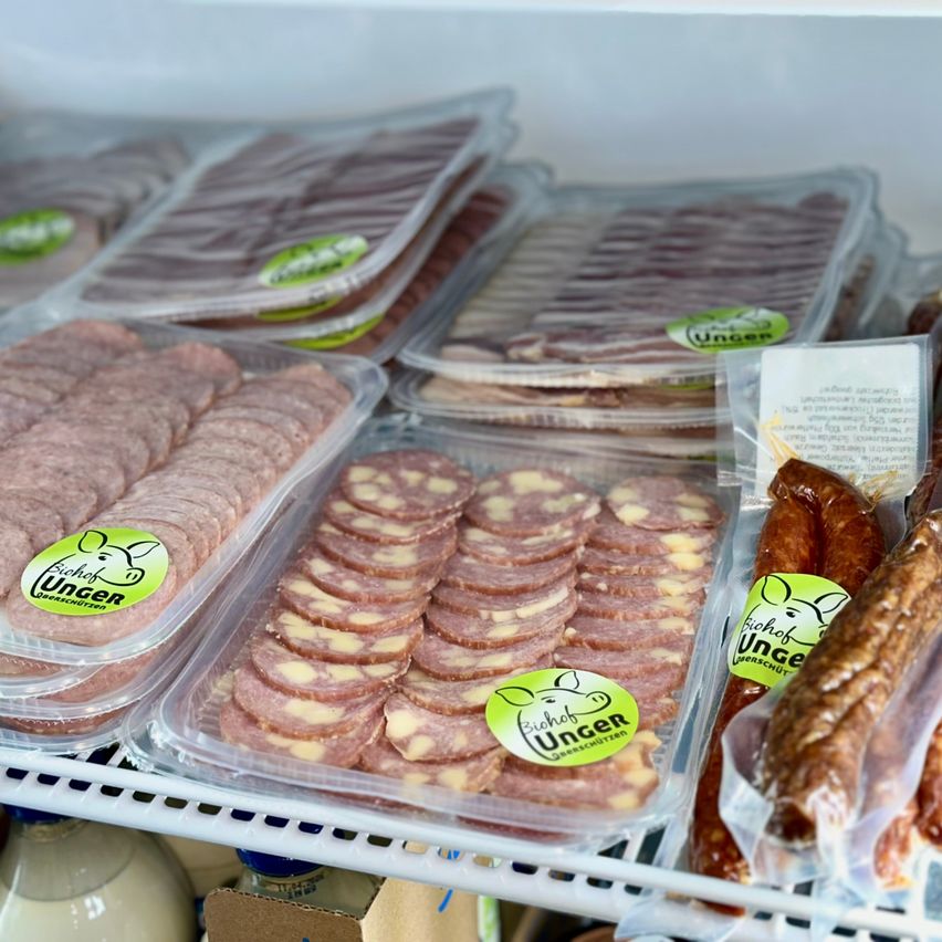 Ein Kühlschrankregal mit verschiedenen verpackten Fleischsorten, einschließlich Würste, Salami und geschnittenem Fleisch, alles in durchsichtigen Plastikbehältern mit grünen Etiketten.