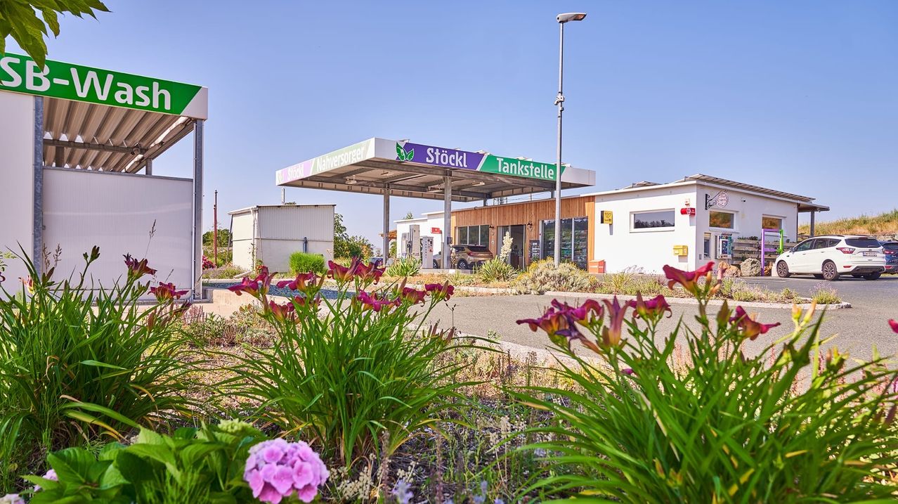 Eine moderne Tankstelle mit einem geparkten Auto, umgeben von Grün und Blumen, unter einem klaren blauen Himmel.