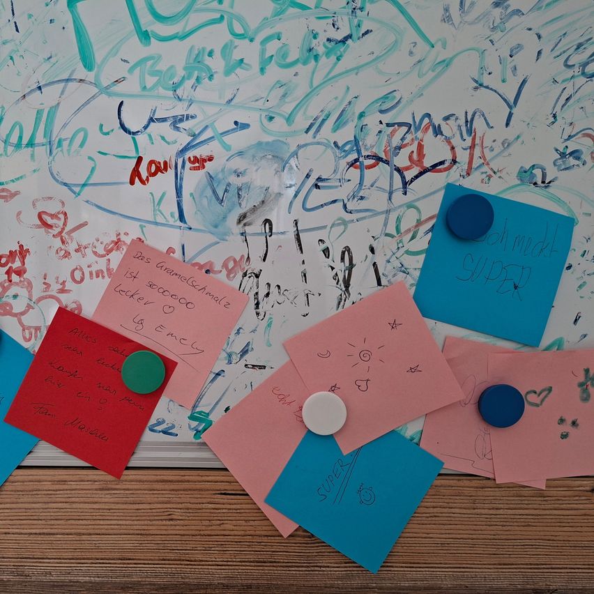 Eine weiße Tafel mit farbenfrohen Zeichnungen und Haftnotizen mit handgeschriebenen Nachrichten. Einige Notizen haben Knöpfe befestigt. Die Tafel steht auf einer Holzoberfläche.