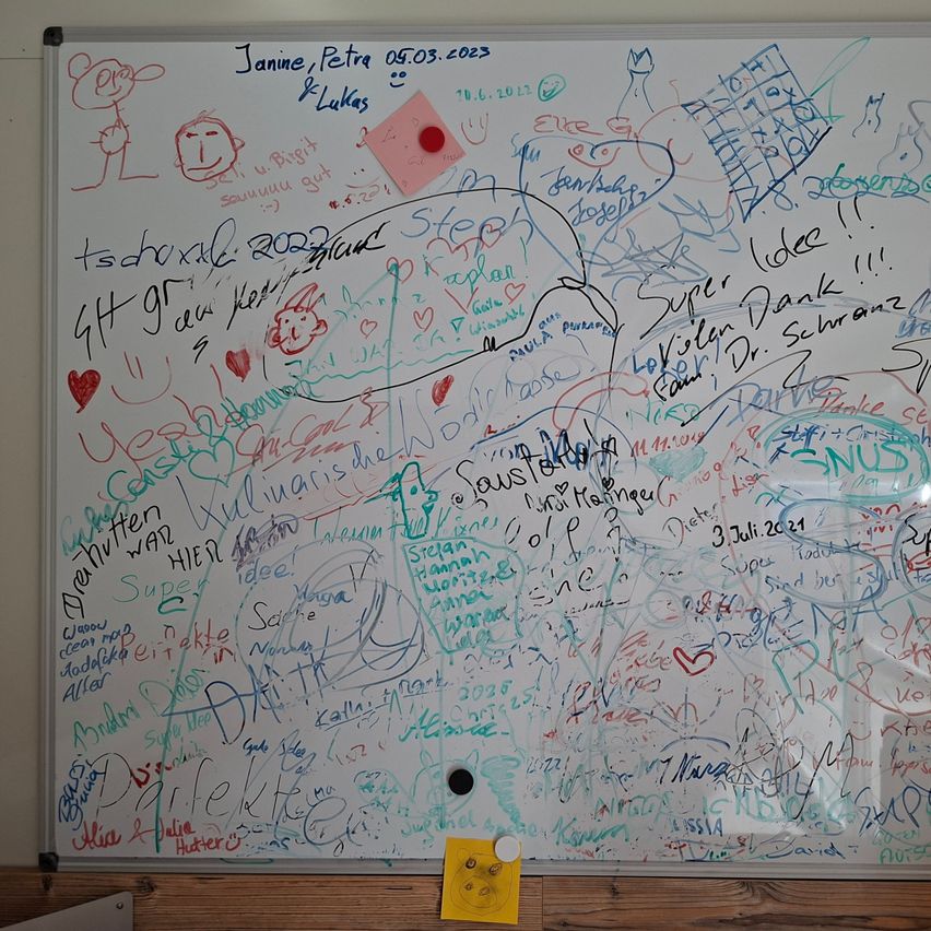 Ein Whiteboard, das mit verschiedenen farbigen Zeichnungen, Unterschriften und Nachrichten bedeckt ist. Es gibt Namen, Daten und einige Illustrationen. Ein roter Knopf ist in der oberen rechten Ecke angeheftet.