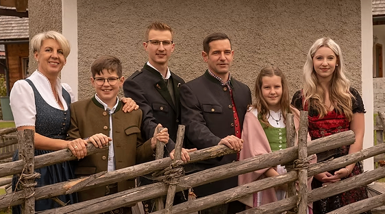 Eine Familie posiert für ein Foto vor einem Gebäude mit einem Holzzaun. Die Familienmitglieder lächeln und tragen traditionelle Kleidung.