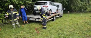 Drei Feuerwehrleute arbeiten an einem Abschleppwagen und ziehen ein silbernes Auto aus einem von dichten Bäumen umgebenen Grasfeld.