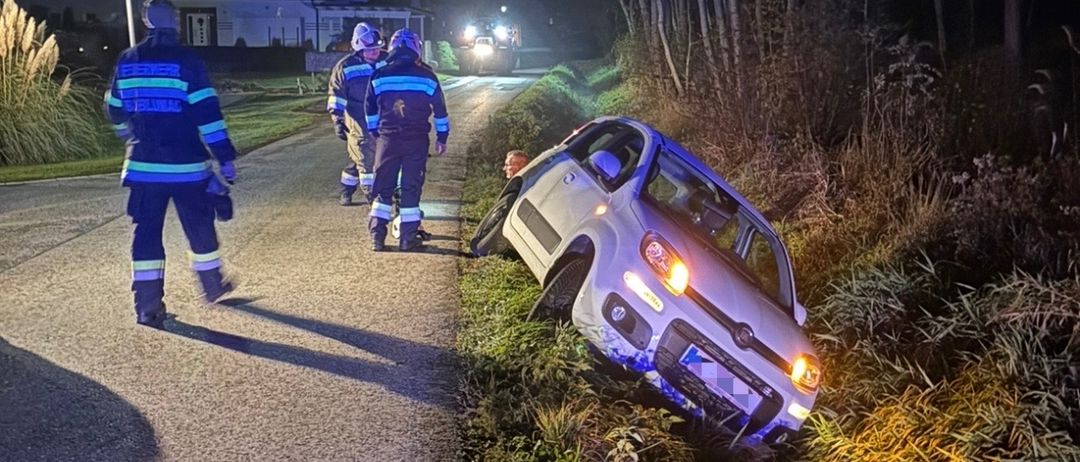 Ein Auto liegt nachts auf dem Landweg auf der Seite, drei Feuerwehrleute sind vor Ort. Das Fahrzeug steht an einem Grasstreifen.