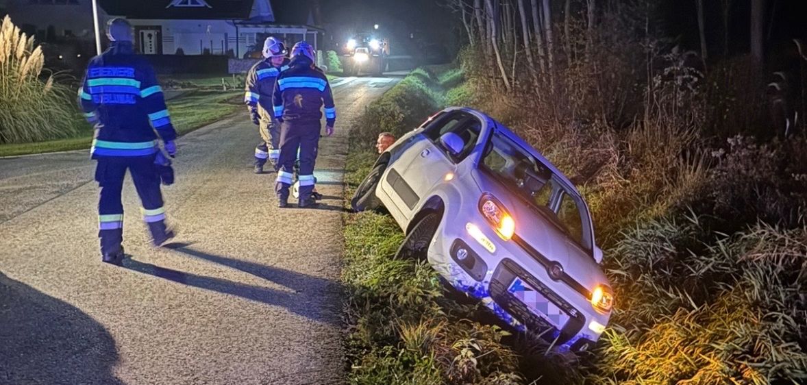 Ein Auto liegt nachts auf dem Landweg auf der Seite, drei Feuerwehrleute sind vor Ort. Das Fahrzeug steht an einem Grasstreifen.