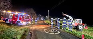 In der Nacht ist ein Feuerwehrwagen am Straßenrand geparkt, umringt von Feuerwehrleuten. Ein roter Traktor steht auf dem Rasen.