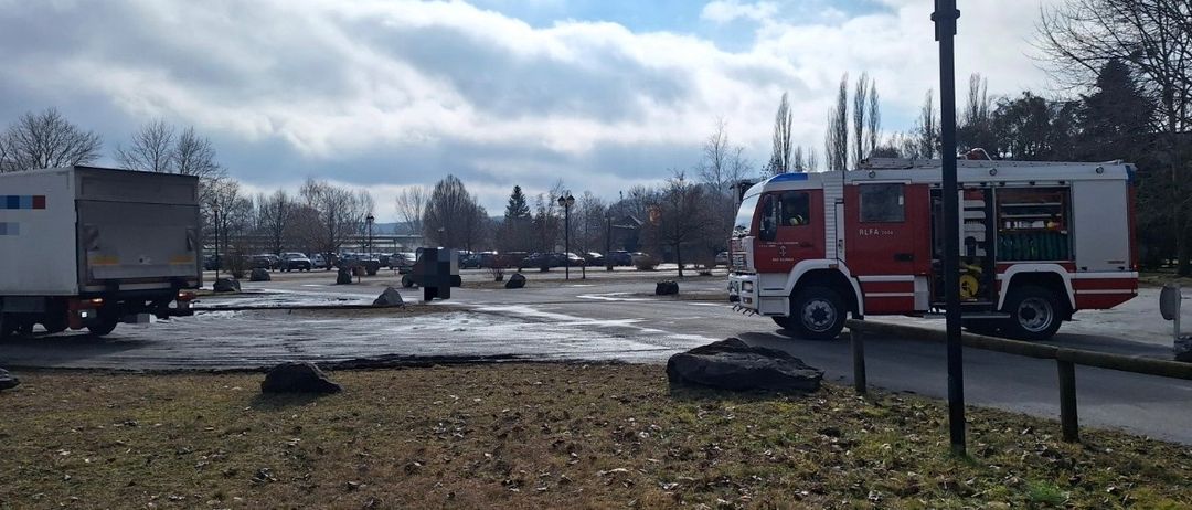 Ein Feuerwehrauto steht auf einem Parkplatz mit einigen Autos. Der Himmel ist bewölkt.