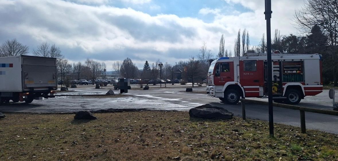 Ein Feuerwehrauto steht auf einem Parkplatz mit einigen Autos. Der Himmel ist bewölkt.