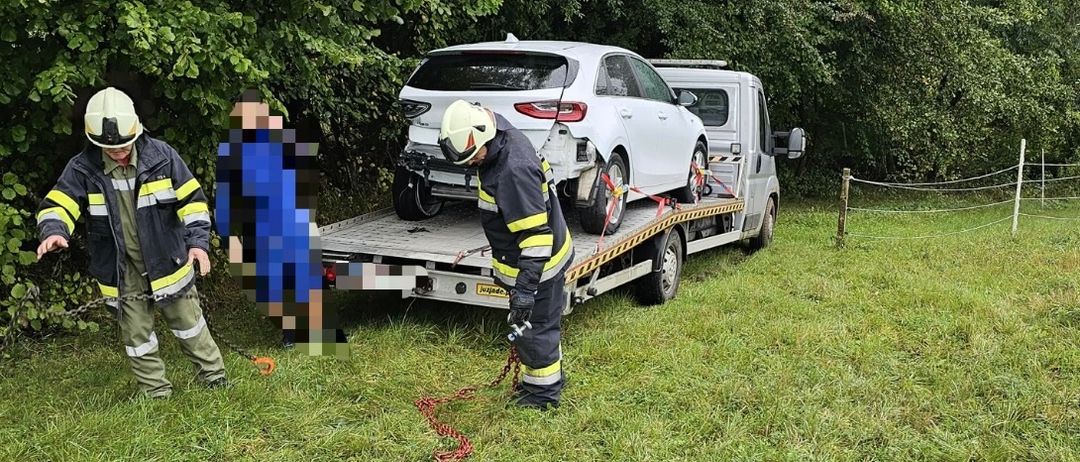 Zwei Feuerwehrleute ziehen ein weißes Auto mit einem Abschleppwagen durch ein von Bäumen umgebenes Grasfeld. Die Vorderseite des Autos ist zur Kamera gerichtet.