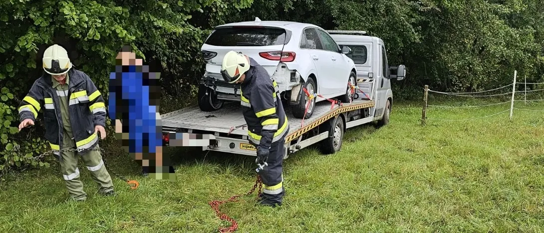Zwei Feuerwehrleute ziehen ein weißes Auto mit einem Abschleppwagen durch ein von Bäumen umgebenes Grasfeld. Die Vorderseite des Autos ist zur Kamera gerichtet.