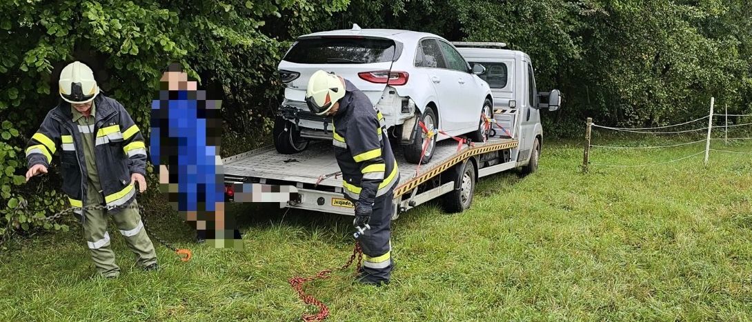 Zwei Feuerwehrleute ziehen ein weißes Auto mit einem Abschleppwagen durch ein von Bäumen umgebenes Grasfeld. Die Vorderseite des Autos ist zur Kamera gerichtet.