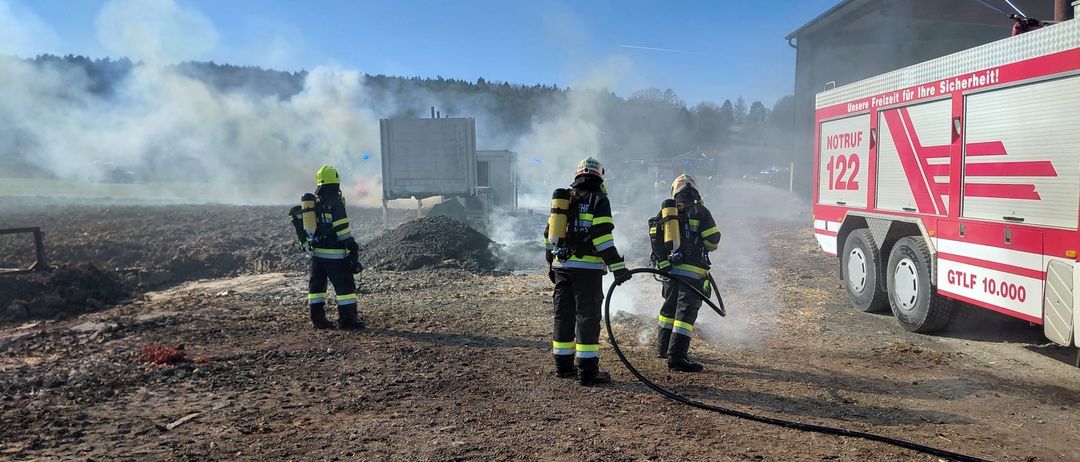 Drei Feuerwehrleute sind draußen und benutzen einen Schlauch, um ein Feuer zu löschen. Sie sind mit Helmen und Schutzanzügen ausgestattet. Ein LKW mit der Nummer 122 ist in der Nähe geparkt.