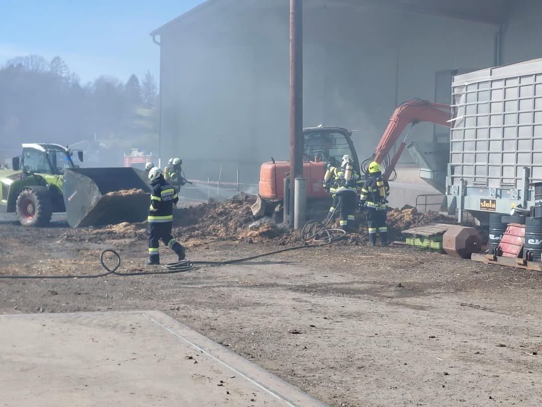 Feuerwehrleute in Schutzkleidung stehen an einem Ort mit starkem Rauch. Sie befinden sich in der Nähe eines Gebäudes und schwerer Maschinen.