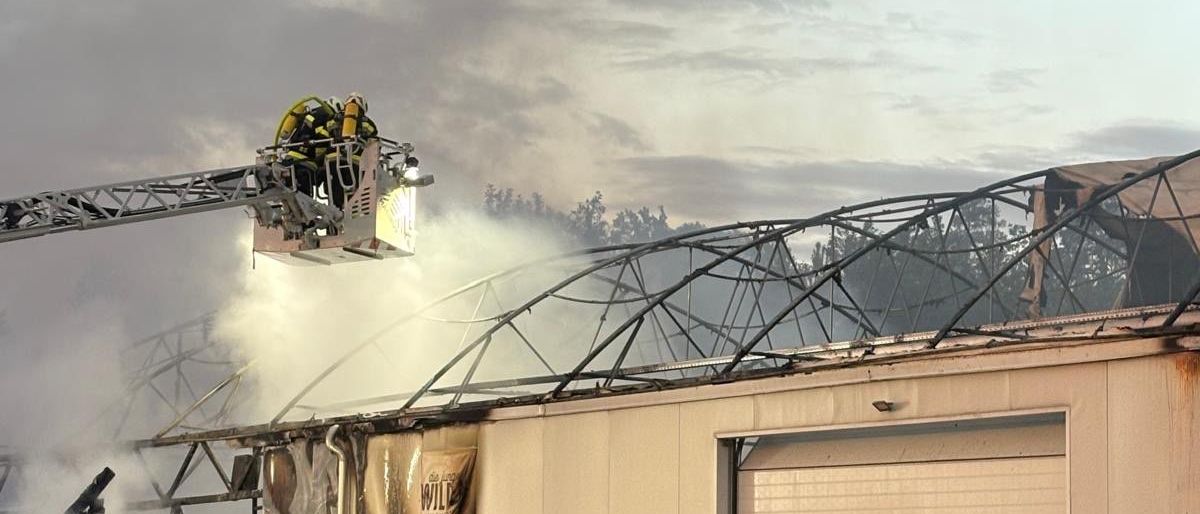 Feuerwehrleute bekämpfen einen Brand in einem Gebäude, einer auf einer Leiter spritzt Wasser. Das Gebäude weist erhebliche Schäden auf und Rauch ist sichtbar.