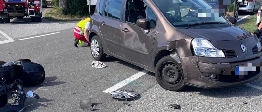 Die Szene eines Autounfalls mit einem beschädigten Auto und Motorradteilen auf der Straße. Eine Person in einer reflektierenden Weste kümmert sich um das Motorrad.