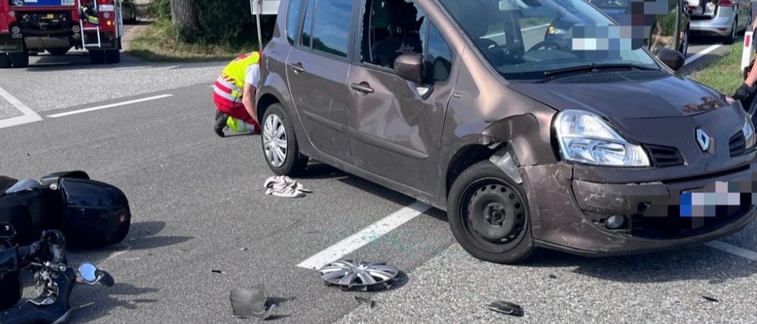 Die Szene eines Autounfalls mit einem beschädigten Auto und Motorradteilen auf der Straße. Eine Person in einer reflektierenden Weste kümmert sich um das Motorrad.