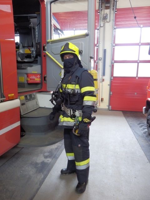 Ein Feuerwehrmann in voller Ausrüstung steht vor einem Feuerwehrwagen, wobei die Tür des Wagens offen ist und die Ausrüstung im Inneren sichtbar ist.
