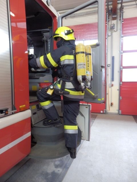 Ein Feuerwehrmann in voller Ausrüstung, einschließlich eines gelben Helms, klettert die Stufen eines Feuerwehrwagens hinauf. Er hält sich mit der rechten Hand an der Seite des Wagens fest.