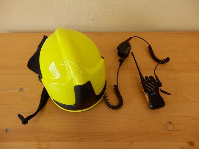 Ein gelber Sicherheitshelm mit schwarzem Riemen und ein Walkie-Talkie mit gewickeltem Kabel liegen auf einer Holzoberfläche.