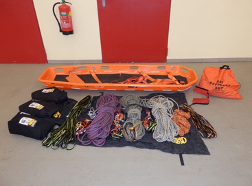 Eine orangefarbene Trage ist auf dem Boden ausgelegt, zusammen mit verschiedenen Seilen, Taschen und Sicherheitsausrüstung. Ein Feuerlöscher ist an der Wand dahinter montiert.