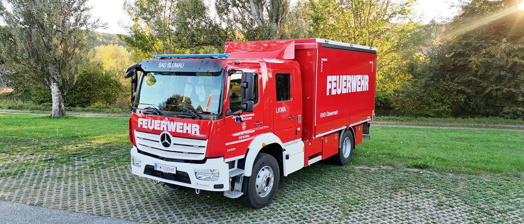 Ein roter Mercedes-Benz Feuerwehrwagen mit 'FEUERWEHR' auf der Seite und 'BAD BLUMA' vorne, geparkt auf einer Kiesfläche mit Bäumen im Hintergrund.